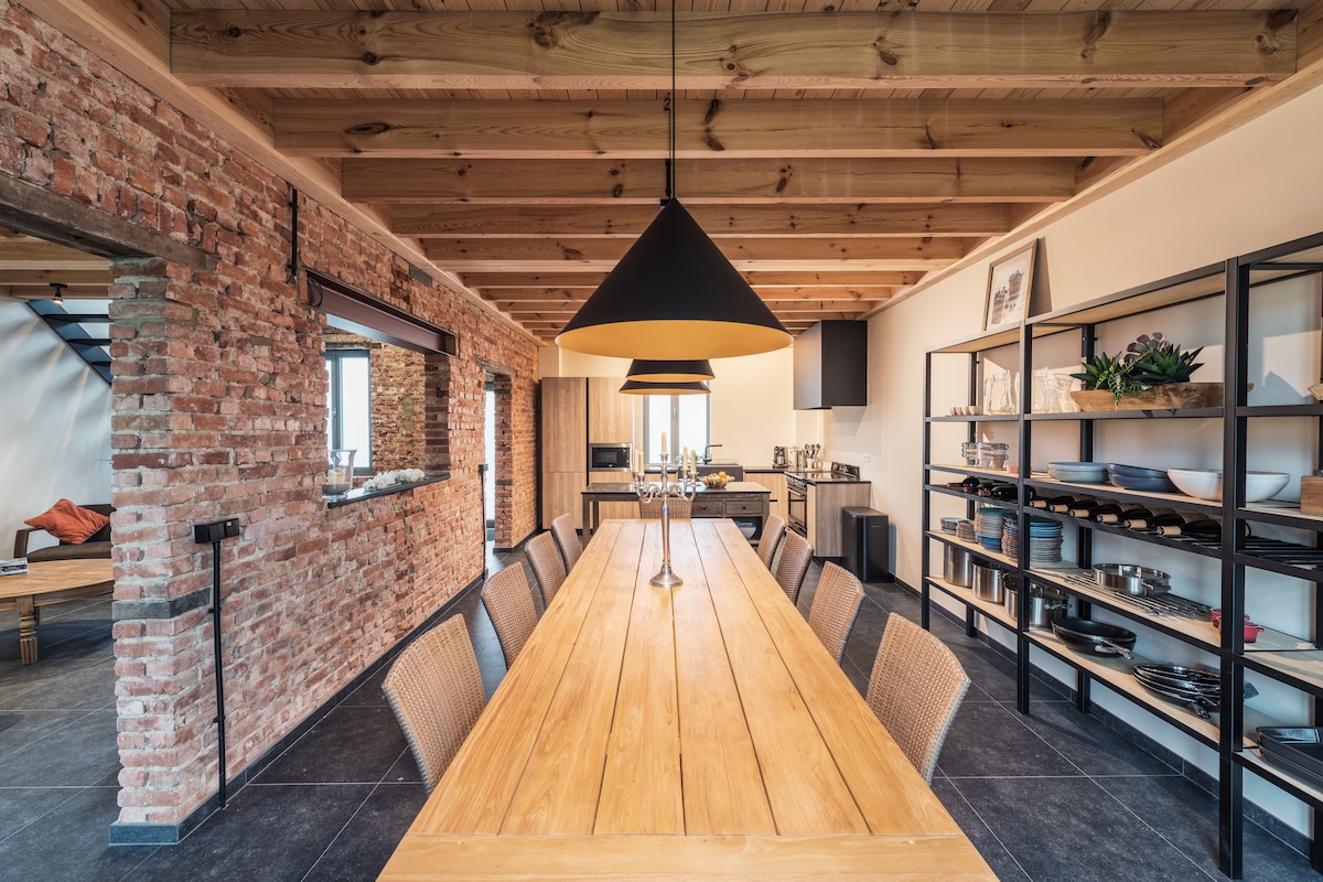 A spacious dining area features a long wooden table surrounded by woven chairs. Exposed wooden beams and brick walls create a rustic ambiance. A dark pendant light hangs above the table, and a neat arrangement of dishes is visible on a nearby shelf.