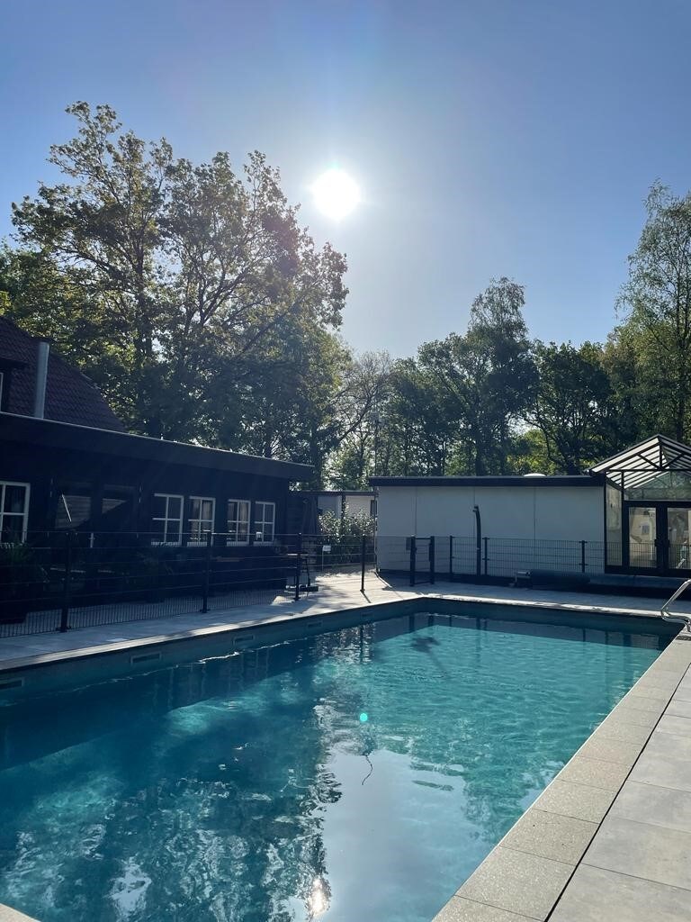 A clear blue sky is seen above a tranquil swimming pool, reflecting sunlight. Surrounding trees provide shade, while a building with large windows is positioned on one side, creating an inviting atmosphere. The pool area is well-maintained with a safety railing for added security.