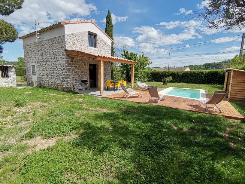 The exterior showcases a charming stone house with a wooden overhang. A private pool is surrounded by a sun deck with lounge chairs, set in a grassy area. Lush greenery frames the scene, while a clear sky adds to the relaxed outdoor environment.