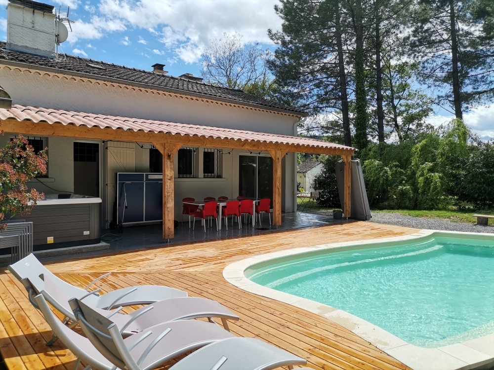An outdoor area features a wooden deck surrounding a clear blue pool. Lounge chairs are arranged nearby, and a shaded dining space with red chairs is situated under a wooden pergola. Lush trees and greenery are visible in the background.