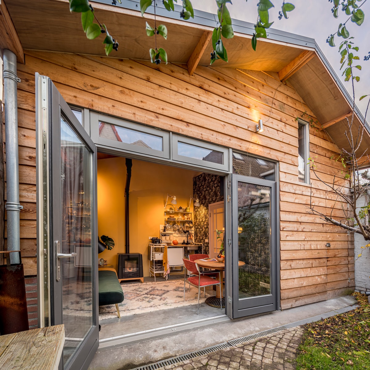 A wooden exterior with large glass doors opens to a cozy interior space. The warm glow from inside highlights a kitchenette, dining area, and comfortable seating. A shared garden is visible through the doors, surrounded by greenery.