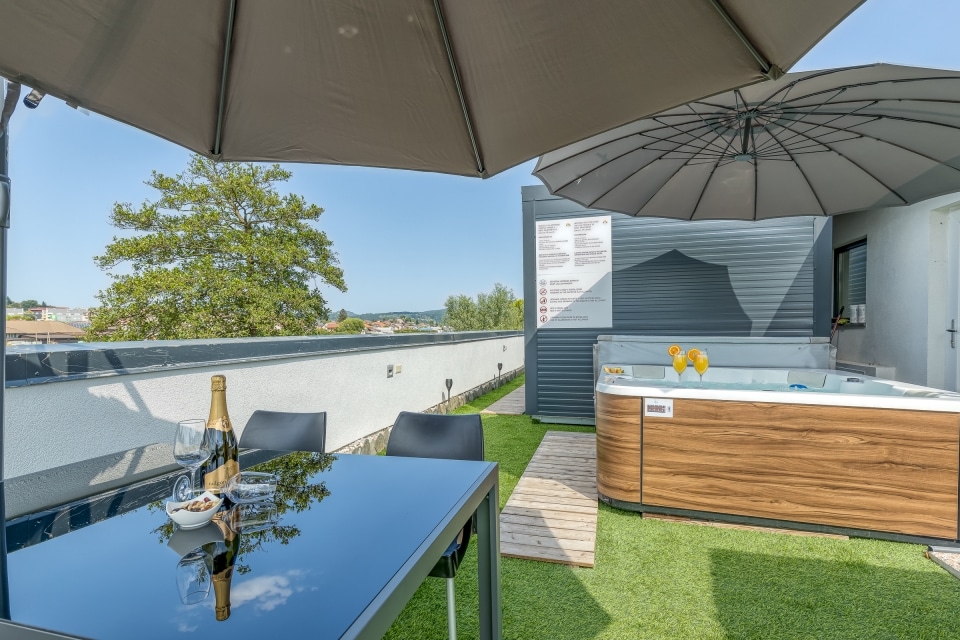 An outdoor terrace features a dining table with glassware and snacks, shaded by large umbrellas. A hot tub is visible, surrounded by a wooden deck and greenery. The expansive view includes distant trees and a clear sky, providing a serene environment for relaxation.