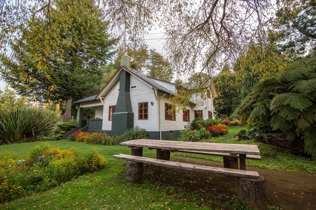 A charming house is nestled among lush greenery, surrounded by vibrant flowerbeds. A wooden picnic table is positioned in front of the house, inviting outdoor relaxation. Large trees provide shade, enhancing the peaceful atmosphere of the serene setting.
