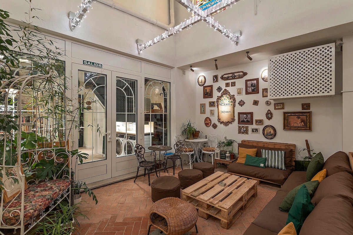 A communal lounge area features a mix of seating options, including a brown sofa adorned with decorative pillows and cozy ottomans. Wall art in various frames decorates the walls, while plants add a touch of greenery. A large glass door leads to an outdoor space.