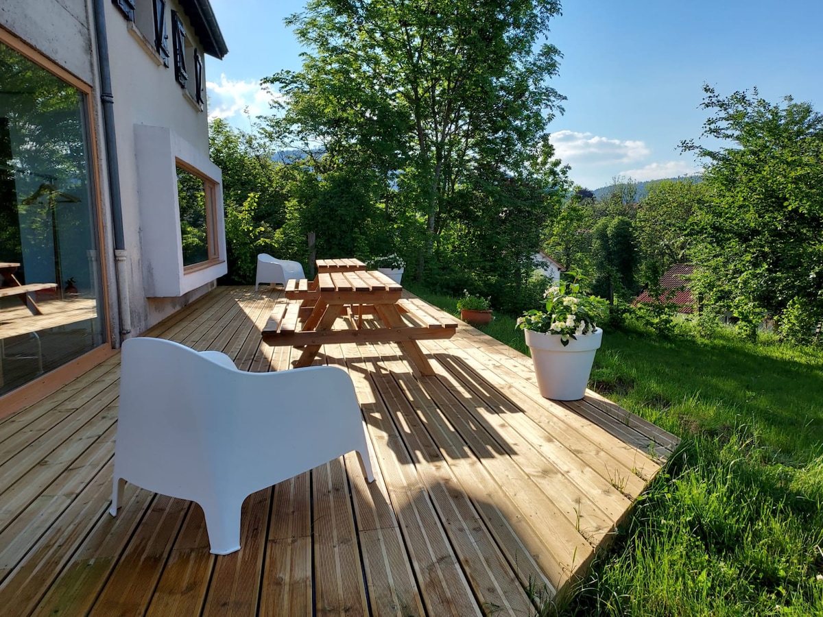 A wooden deck is presented with a large picnic table surrounded by white outdoor chairs. Lush green grass and trees are visible in the background, complemented by a clear sky. A potted plant adds a touch of greenery to the outdoor space.