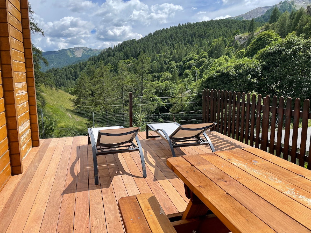 A spacious wooden terrace features two lounge chairs positioned under natural light. A wooden dining table is visible, providing a place to gather. Surrounded by greenery, the area offers a scenic view of the mountains and trees, enhancing the outdoor experience.
