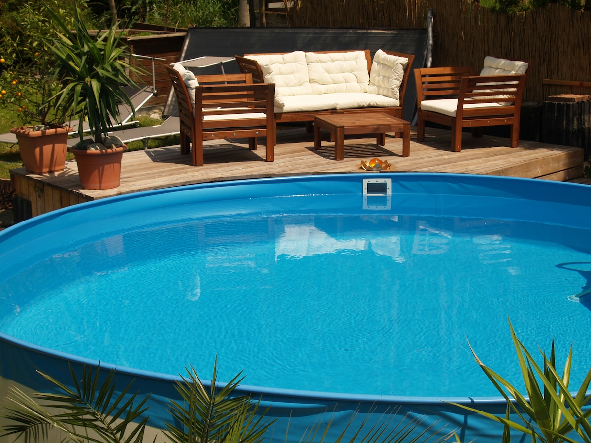 A circular blue pool is positioned at the center, surrounded by wooden decking. Comfortable seating is arranged nearby, with plush cushions on a sofa and an accompanying table. Lush green plants are present, enhancing the refreshing outdoor space.