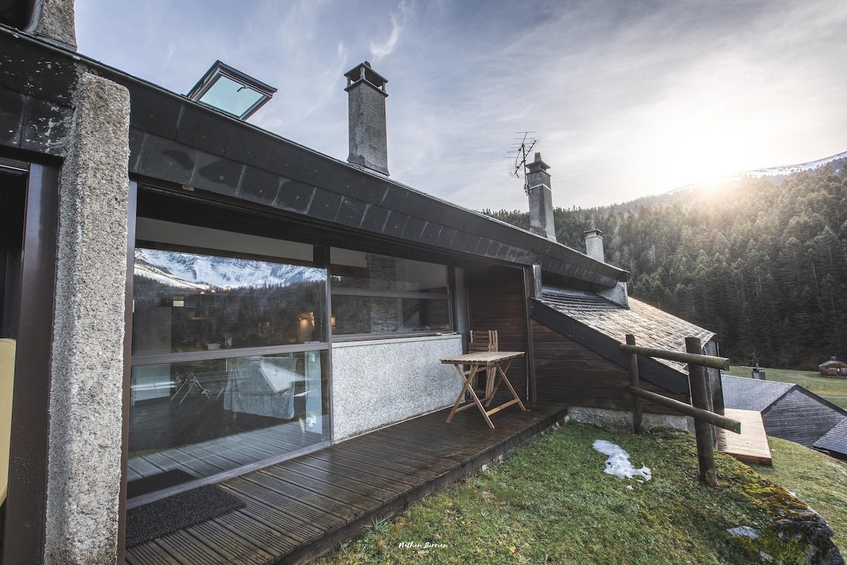 The exterior of the gîte is shown, featuring a wooden terrace with a small table and chair. Smoke rises gently from the chimney, and the surrounding landscape consists of lush greenery and mountains, creating a serene atmosphere. Soft sunlight filters through the clouds, illuminating the scene.