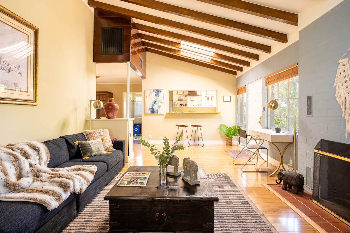 A spacious living area features a cozy dark sofa adorned with a soft throw. Exposed wooden beams span the ceiling, while natural light filters through large windows, illuminating a coffee table and a small dining area in the background.