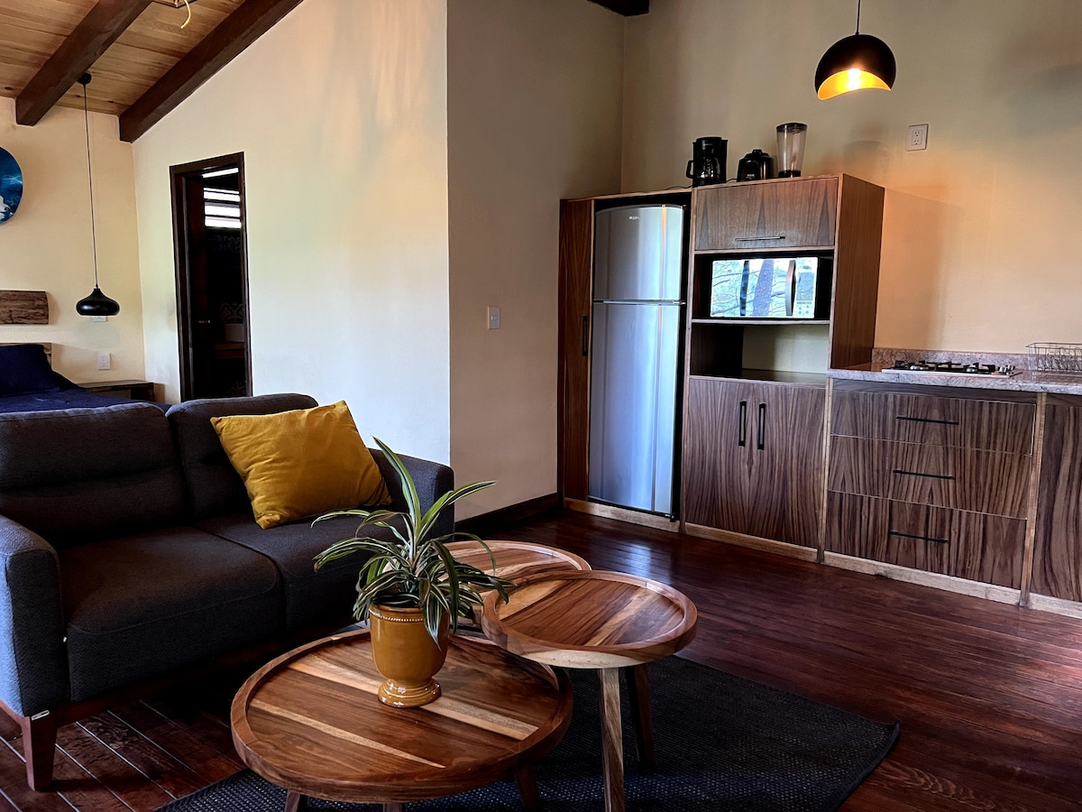 A light-filled living space features a comfortable gray sofa adorned with a yellow pillow. Two wooden tables are positioned in front of the sofa, alongside a potted plant. The kitchen area is equipped with modern appliances, including a refrigerator and a stove, offering a functional layout.