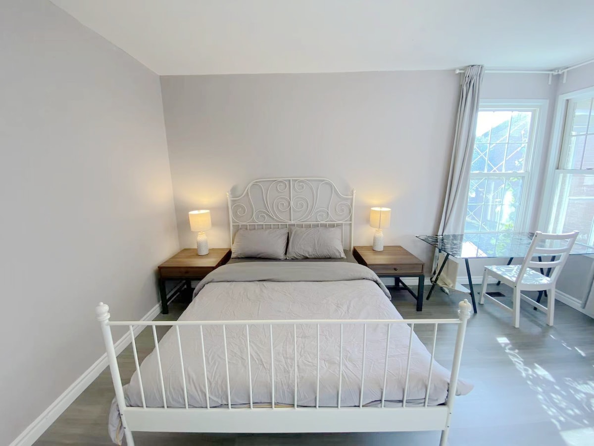 The room features a white metal bed with a plush bedding set and two bedside lamps on wooden tables. A clear glass desk is positioned beside a window, allowing natural light to illuminate the space. A simple chair complements the desk, offering a cozy workspace.