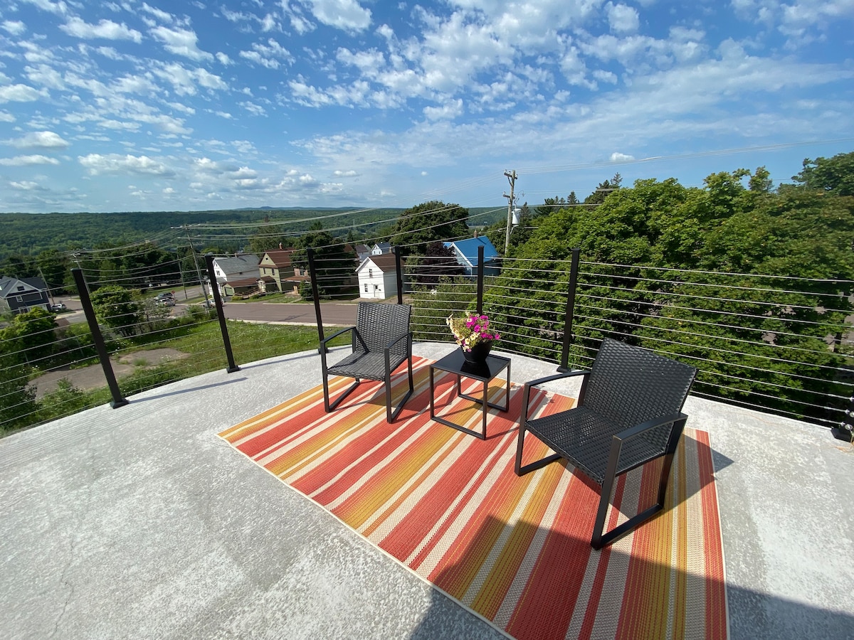 A spacious outdoor patio area features three chairs arranged around a small table, adorned with a flower centerpiece. A striped outdoor rug adds color beneath them, while scenic views of greenery and blue skies create a serene environment.