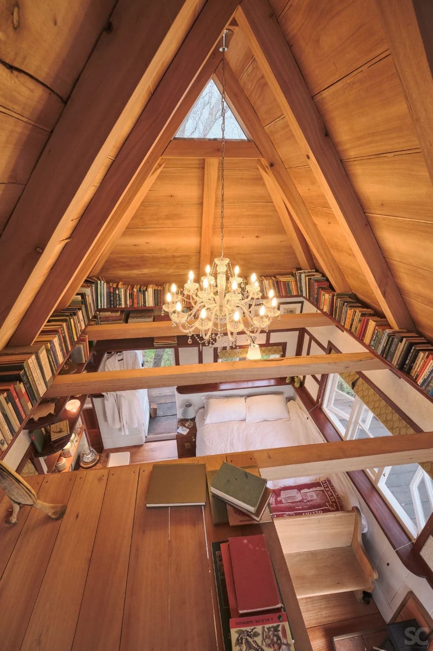A view from above showcases the triangular wooden structure adorned with a chandelier. Shelves filled with books line the perimeter, framing the cozy sleeping area below. Natural light streams in through large windows, highlighting the craftsmanship of the wooden beams and warm tones.