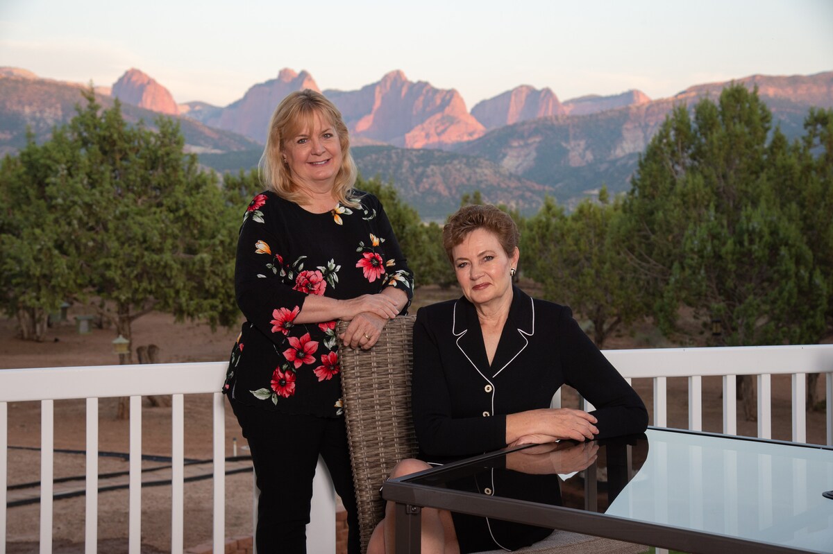Two individuals are captured on a deck overlooking a stunning mountain landscape. A spacious table is set in front, with lush trees framing the view. The warm tones of the mountains contrast with the clear sky, creating a serene backdrop for outdoor relaxation.