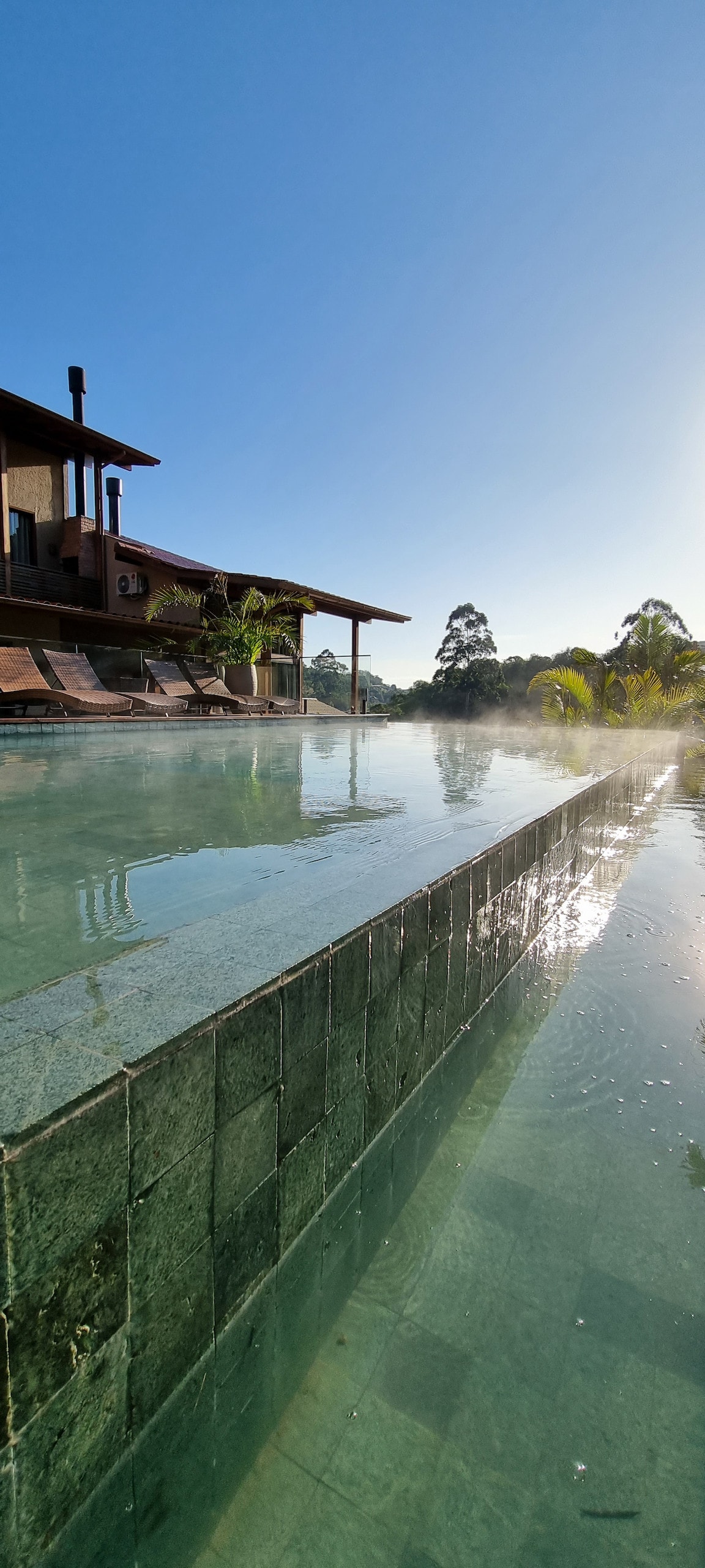 A tranquil swimming pool stretches across the image, featuring a glassy surface reflecting sunlight. The pool is bordered by a stone wall, and lounge chairs are positioned nearby under the shade of a spacious roof. Lush greenery surrounds the area, enhancing the serene environment.