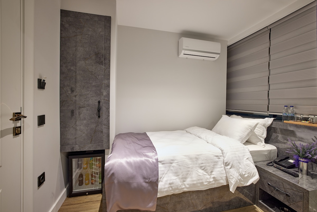 A compact hotel room features a spacious bed with crisp white linens, complemented by a lavender throw at the foot. A small refrigerator is visible beside the bed, and two water bottles are placed on the nightstand, with a textured wall behind the headboard.