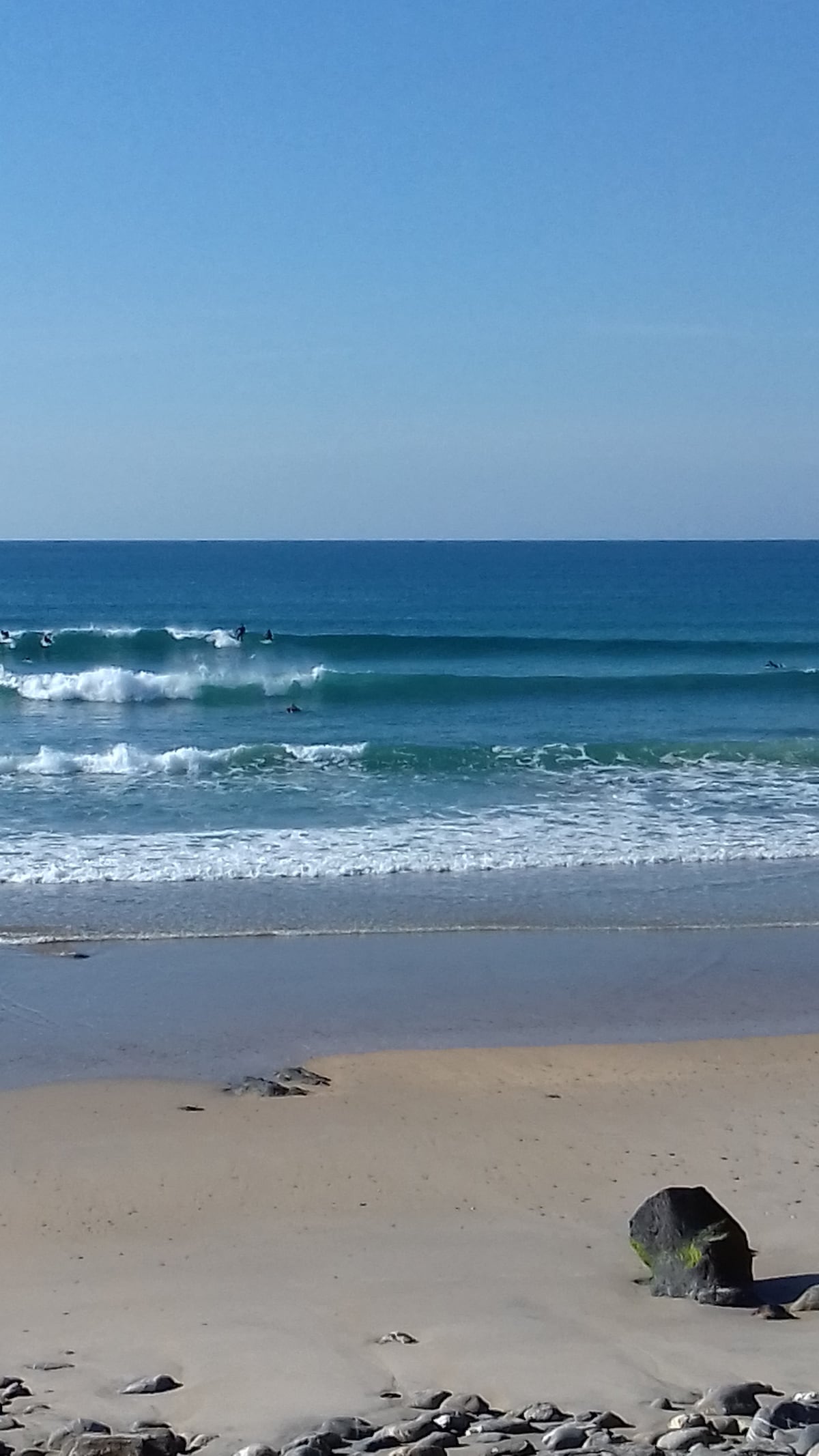 A serene beach scene features gentle waves rolling onto the sandy shore. Surfboard riders can be seen enjoying the ocean, while a rocky formation is present in the foreground, adding texture to the coastal landscape. The sky is clear, providing a bright backdrop.
