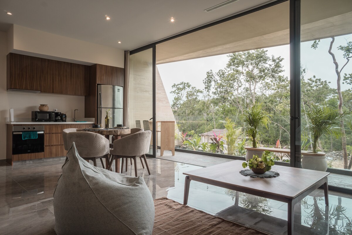 A modern living space is presented with large glass doors offering views of the lush greenery outside. The area features a dining table with four upholstered chairs, a cozy bean bag, and a wooden coffee table topped with fruit. The open kitchen is partially visible.
