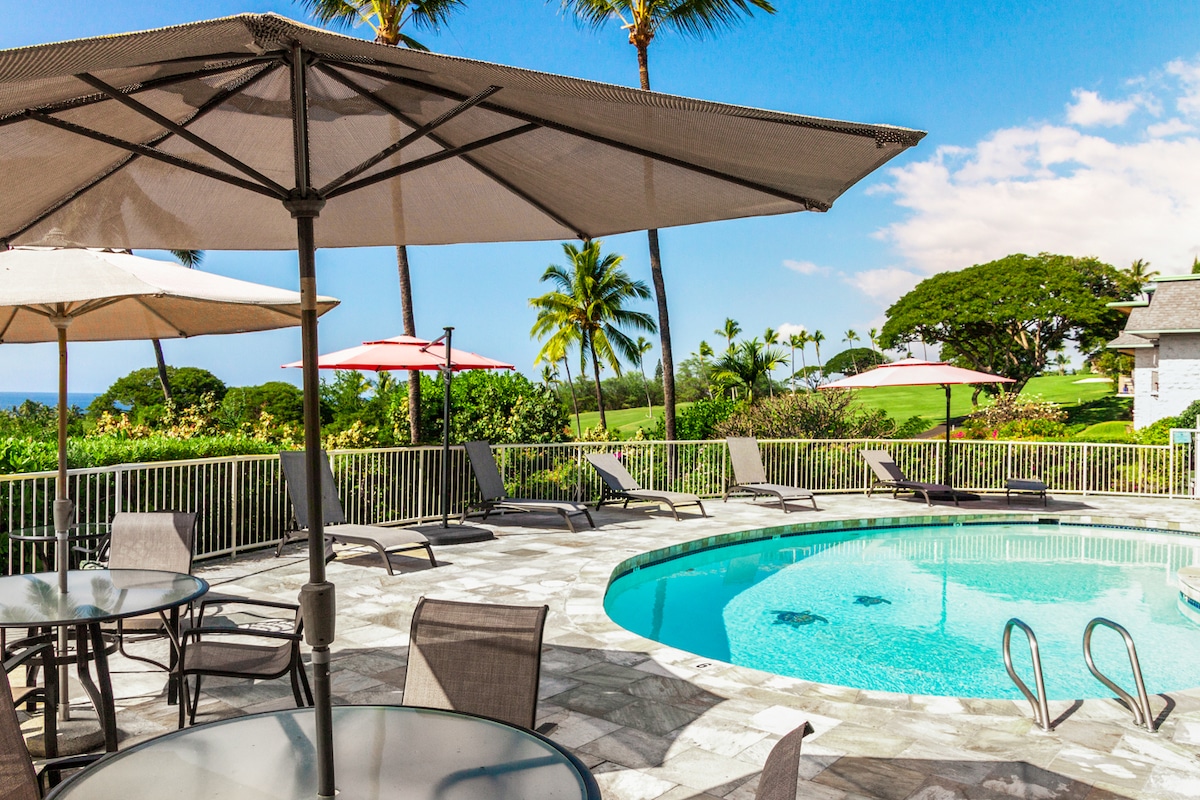 A salt-water pool area is surrounded by a spacious stone deck, featuring several tables with umbrellas. Palm trees frame the scene, with fairway views visible in the background under a clear blue sky.