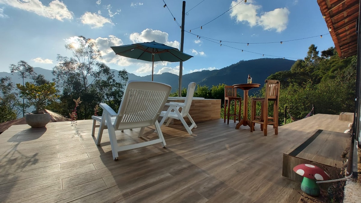 A spacious wooden deck is presented with two white lounge chairs and a small bistro table with two high chairs. An umbrella provides shade, and string lights add ambient lighting. Scenic mountain views are visible in the background, with lush greenery surrounding the area.