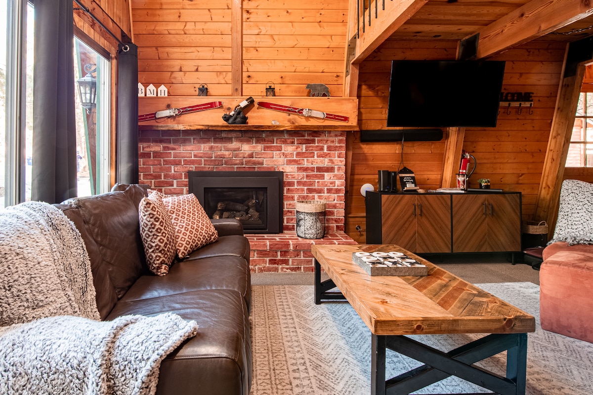 The living area features warm wooden walls and an inviting fireplace surrounded by brick. A spacious brown leather sofa is adorned with patterned throw pillows. A rustic coffee table sits in the center, with a patterned area rug beneath it, enhancing the cozy ambiance.