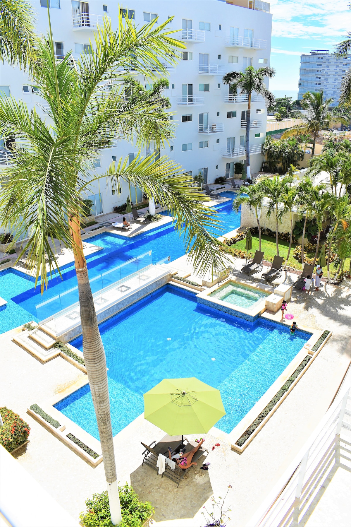 A vibrant pool area is surrounded by lush palm trees and manicured landscaping. Multiple swimming pools of varying sizes are visible, alongside inviting lounge chairs and umbrellas. The modern building provides a spacious feel, with light reflecting off the water in the sunny atmosphere.