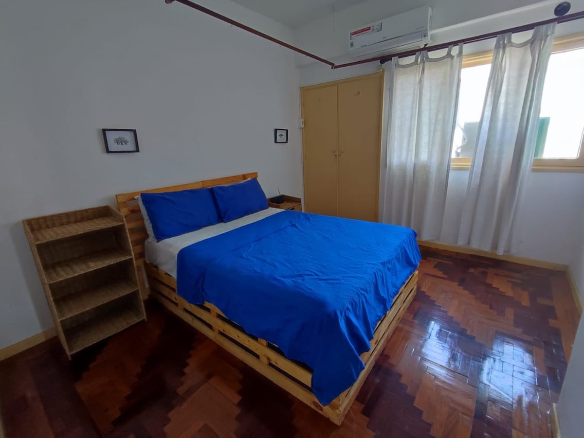 A well-lit bedroom features a wooden bed with a blue bedspread, complemented by matching pillows. A tall, open shelving unit stands nearby, providing storage. Natural light enters through a window covered with sheer curtains, and light hardwood floors enhance the room's brightness.