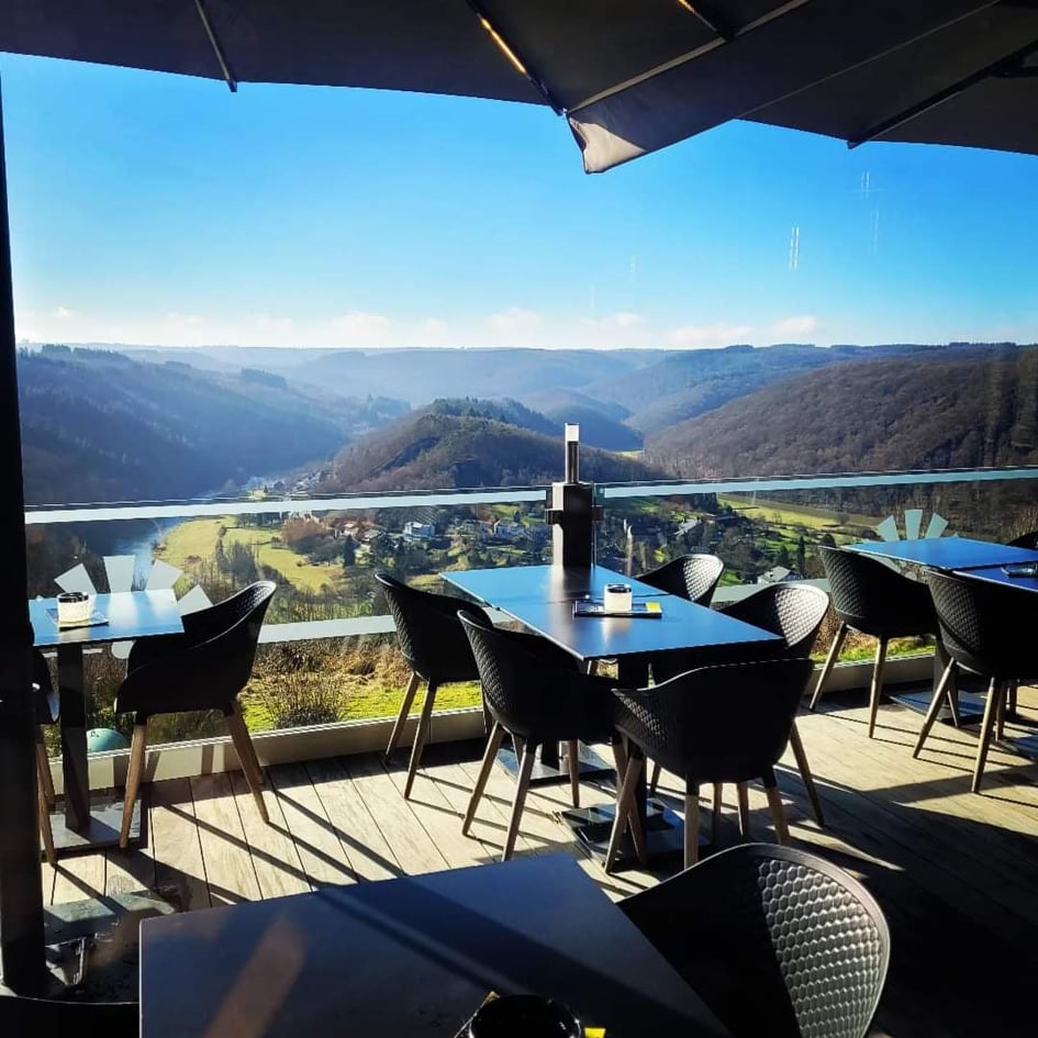 An outdoor dining area features sleek black chairs and tables arranged beside a glass railing. A panoramic view of the valley and hills under a clear blue sky is visible in the background, creating a spacious and open atmosphere.