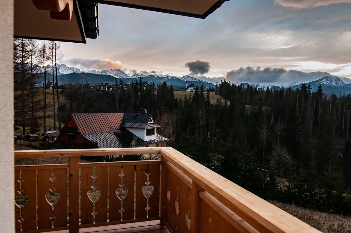 A wooden balcony features ornate railings with heart motifs, overlooking rolling hills and a scenic view of the Tatra Mountains. The landscape transitions from dense evergreen trees to a distant mountain range, with a few residential buildings visible among the greenery.