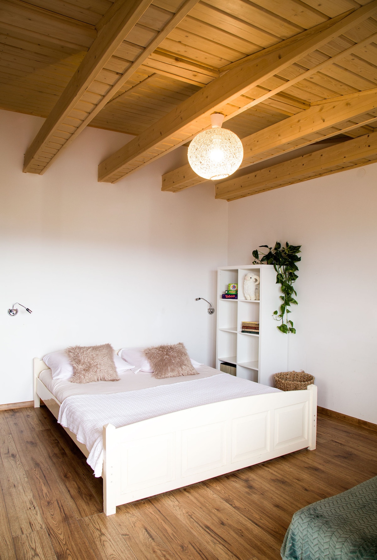 A bright and airy bedroom features a spacious bed with soft pillows, complemented by natural wooden beams across the ceiling. A white shelving unit houses decorative items and books, while a green plant adds a touch of nature to the serene atmosphere.