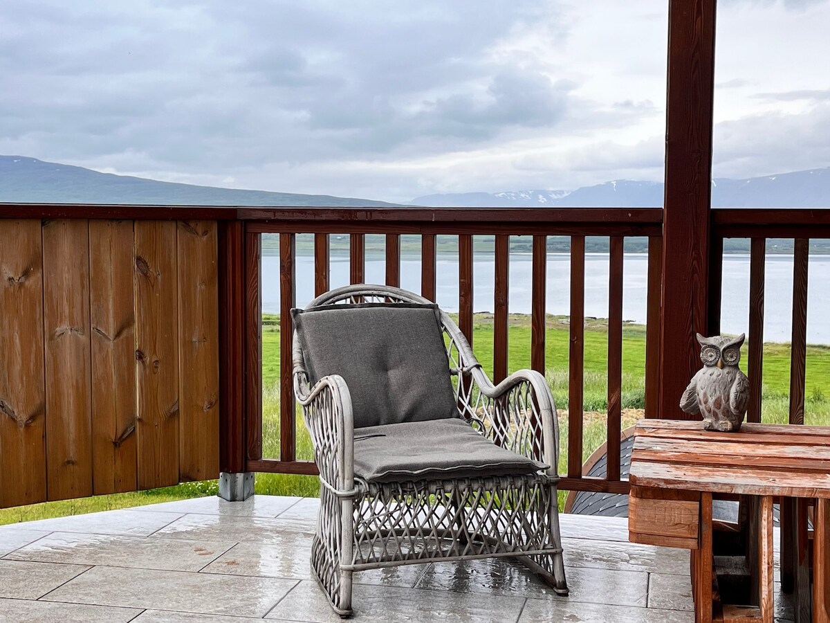 An outdoor seating area features a wicker chair with a soft gray cushion, positioned on a stone patio. A wooden table stands nearby, accompanied by a decorative owl sculpture. The view overlooks a tranquil lake and green landscape under a cloudy sky.