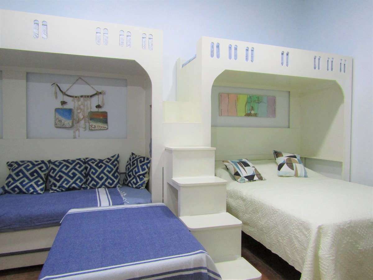 A neat sleeping area features built-in bunk beds with a cozy double bed on the bottom and a single bed on top. Decorative throw pillows add color to the space, complemented by light blue walls and a quilted bedspread.