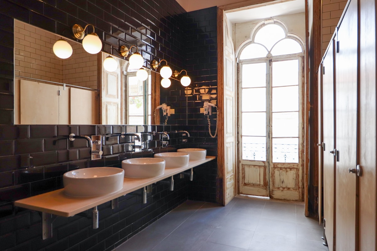A modern bathroom features three round sinks set on a long countertop, illuminated by stylish globe lights. Dark tiles create a sleek look, and two wooden doors lead to private stalls. A large window provides natural light, framed by old wooden shutters.