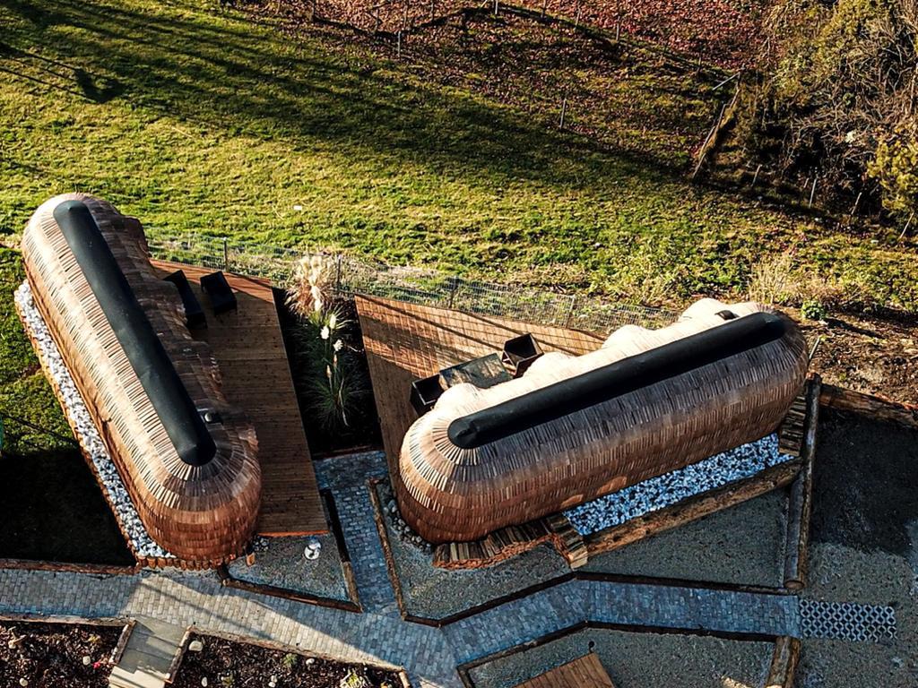 An aerial view reveals two uniquely shaped igloo huts nestled in a green landscape. Surrounding wooden decking and pathways accentuate the natural setting, while spacious outdoor seating areas invite relaxation. The combination of earthy materials and design creates a harmonious retreat in a tranquil environment.