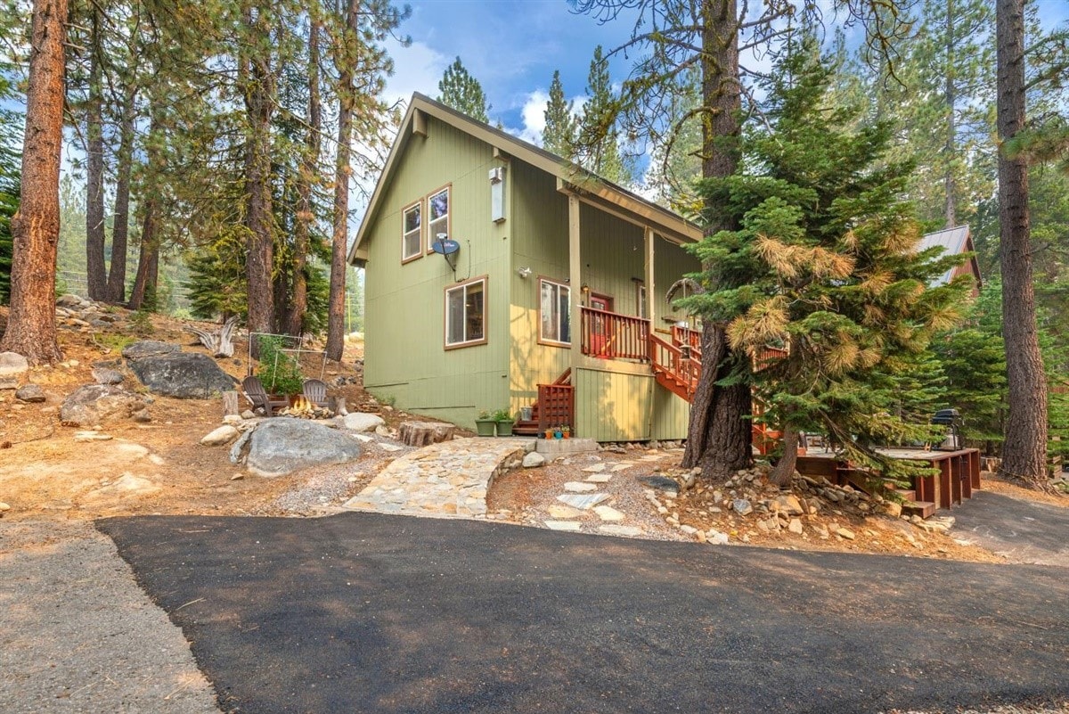 A two-story chalet is nestled among tall pine trees, with a stone pathway leading to the entrance. The exterior features a green facade, complemented by red accents on the porch and railings. Surrounding rocky and wooded areas enhance the natural setting.