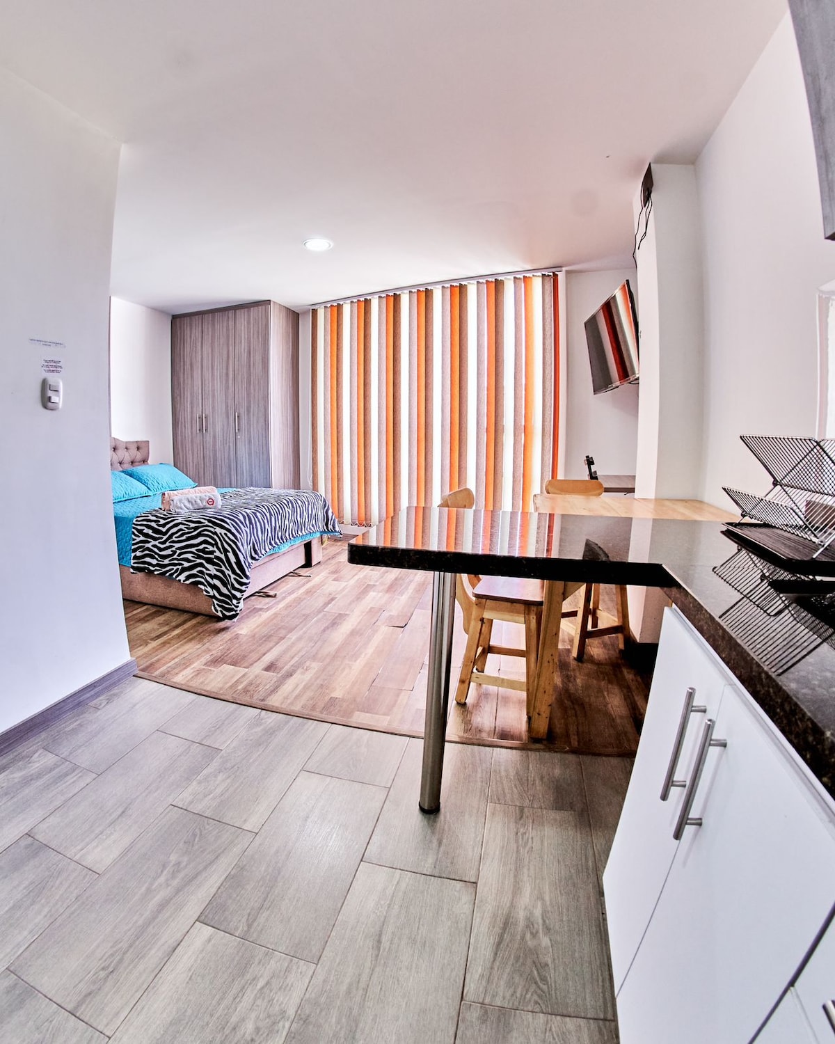 A compact studio apartment features a spacious bed adorned with a striped blanket and a wooden dining table with two chairs. Natural light enters through large, vertical striped curtains, highlighting the warm wooden flooring and modern decor. A wall-mounted television is also visible.