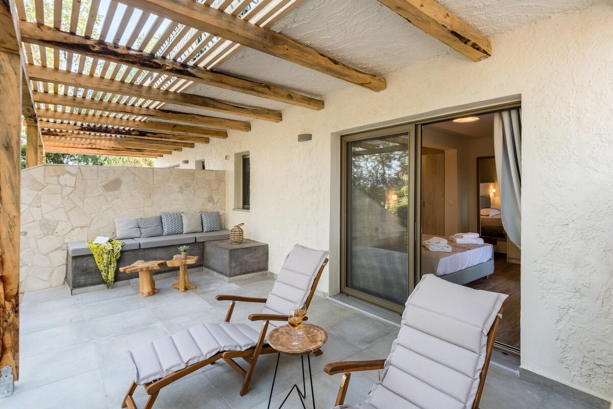 An outdoor terrace is shown, featuring two lounge chairs and a small table. A seating area with cushions and a side table is positioned against a stone wall. Glass doors offer a view into the suite, which is well-lit and furnished.