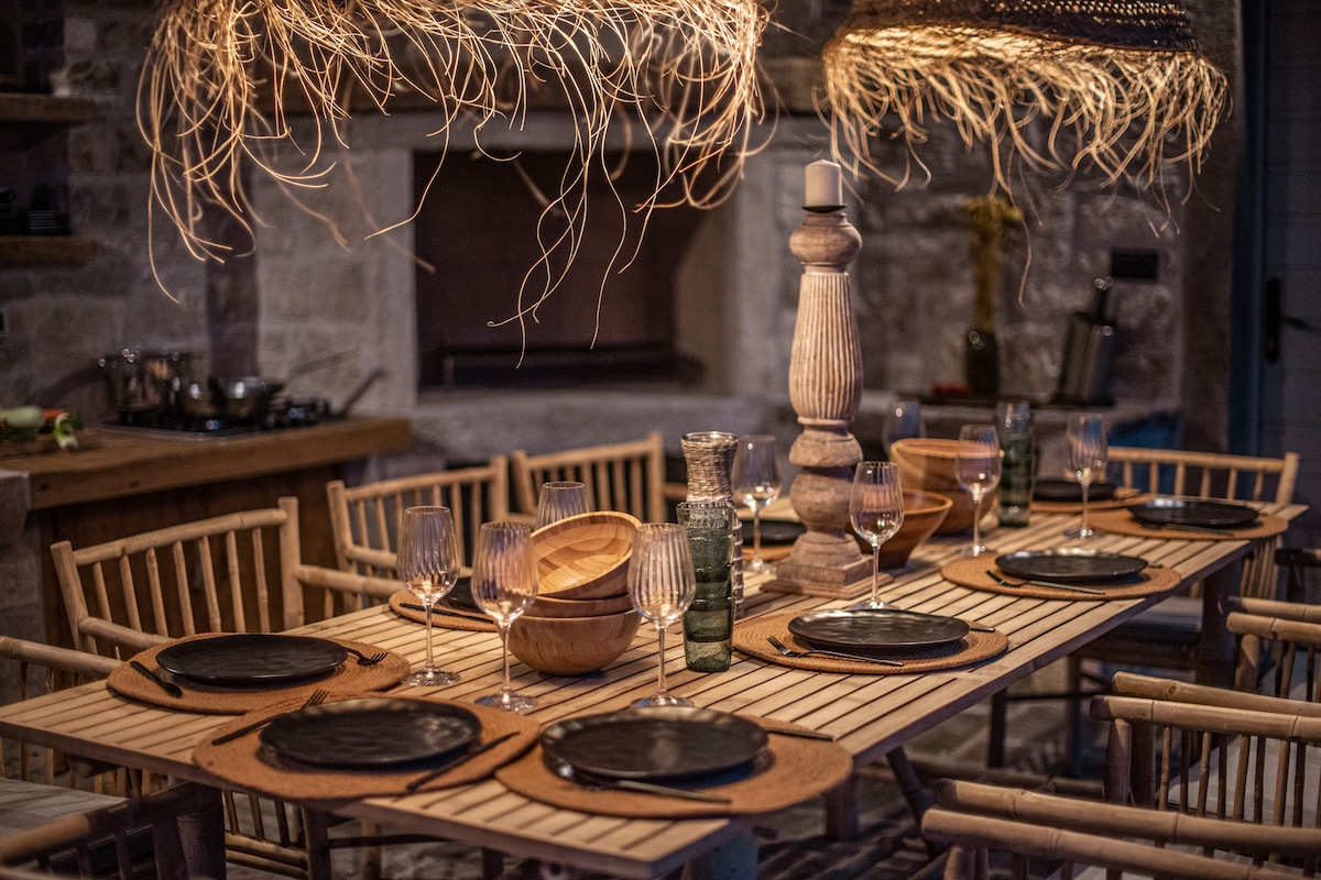 A wooden dining table is set with black plates and glassware, surrounded by natural bamboo chairs. Two large woven light fixtures hang overhead, casting a soft glow. The rustic ambiance is complemented by a stone fireplace in the background and wooden bowls arranged on the table.