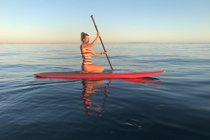 A paddle board is peacefully gliding across calm waters, with a person kneeling at the center. The serene surface reflects the soft colors of the sky during sunset, creating a tranquil scene. The individual is dressed in a striped bathing suit, focusing on the rhythmic motion of paddling.