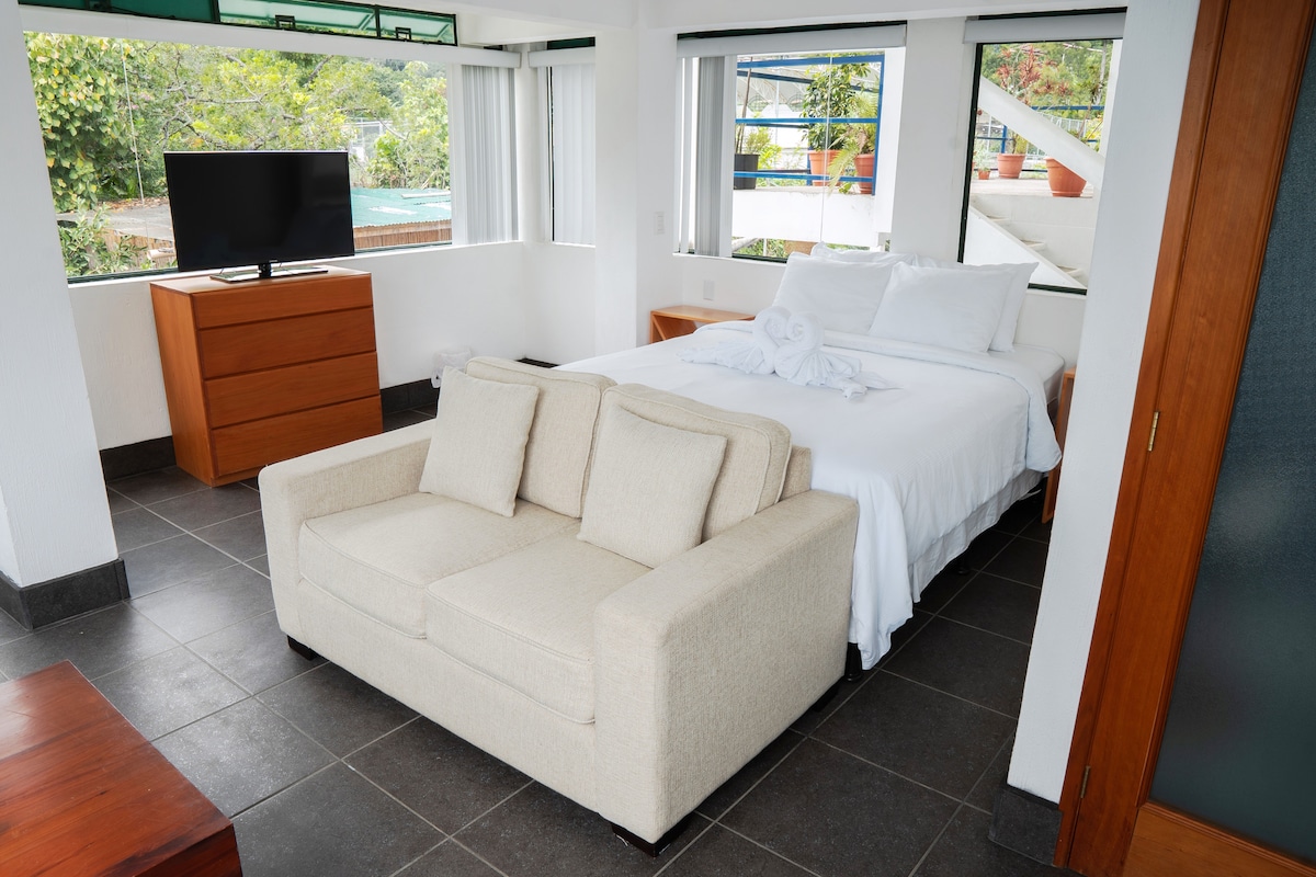 A bright room features a spacious bed dressed in white linens and flanked by large windows that allow natural light to fill the space. A cozy beige sofa sits at the foot of the bed, complemented by wooden furniture including a TV stand.