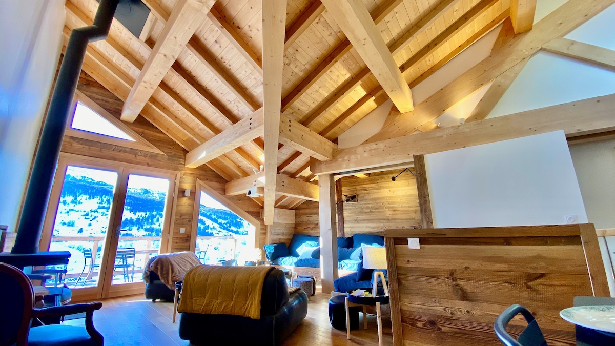 The spacious living area features high, sloped wooden ceilings, with large windows offering natural light and views of the surrounding landscape. Comfortable seating arrangements are set against wooden walls, complemented by a stove for warmth. A dining area is visible, inviting relaxation and gatherings.