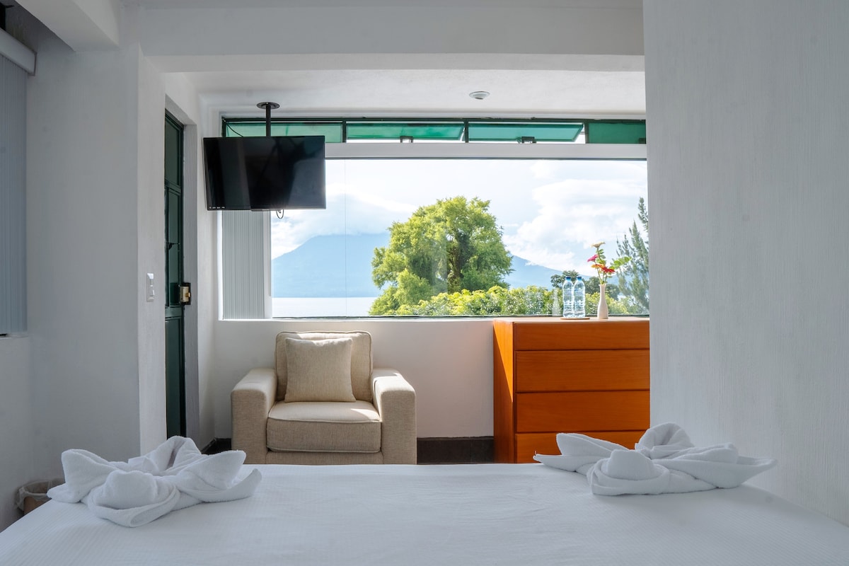 A well-lit room features a spacious bed adorned with neatly arranged towels, set against a backdrop of large windows offering stunning views of the lake and mountains. A cozy armchair is positioned next to a wooden dresser, creating a comforting space for relaxation.