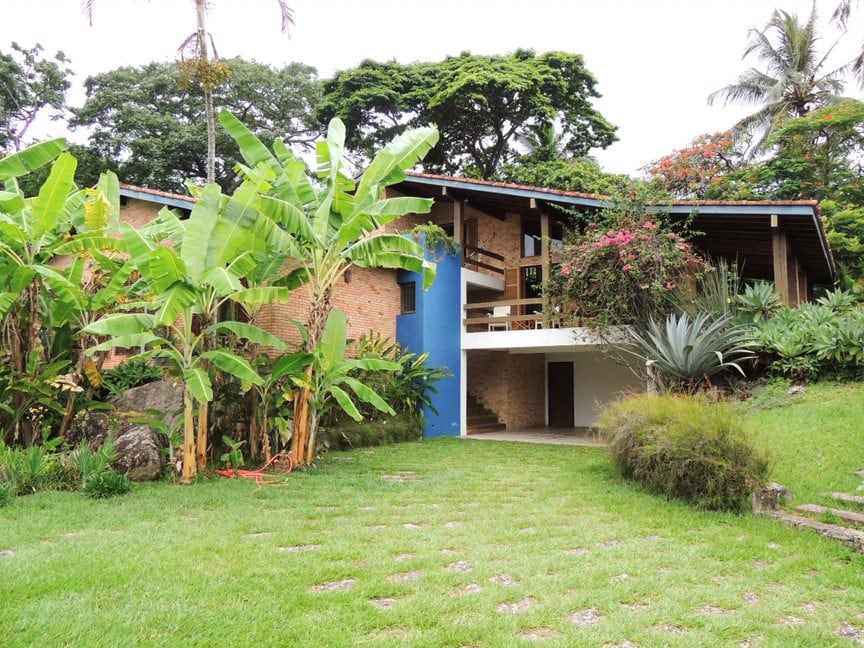 The exterior of the house is surrounded by lush greenery, featuring tall banana plants and other tropical foliage. A pathway lined with stones leads to the entrance, which is accented by vibrant flora. The facade combines earthy tones with a striking blue door.