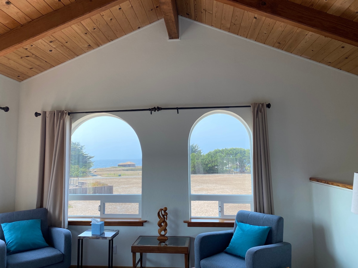 A cozy sitting area features two upholstered chairs with blue cushions, positioned beside a small table. Arched windows offer views of the serene landscape and ocean, while soft natural light filters through sheer curtains, enhancing the inviting ambiance.
