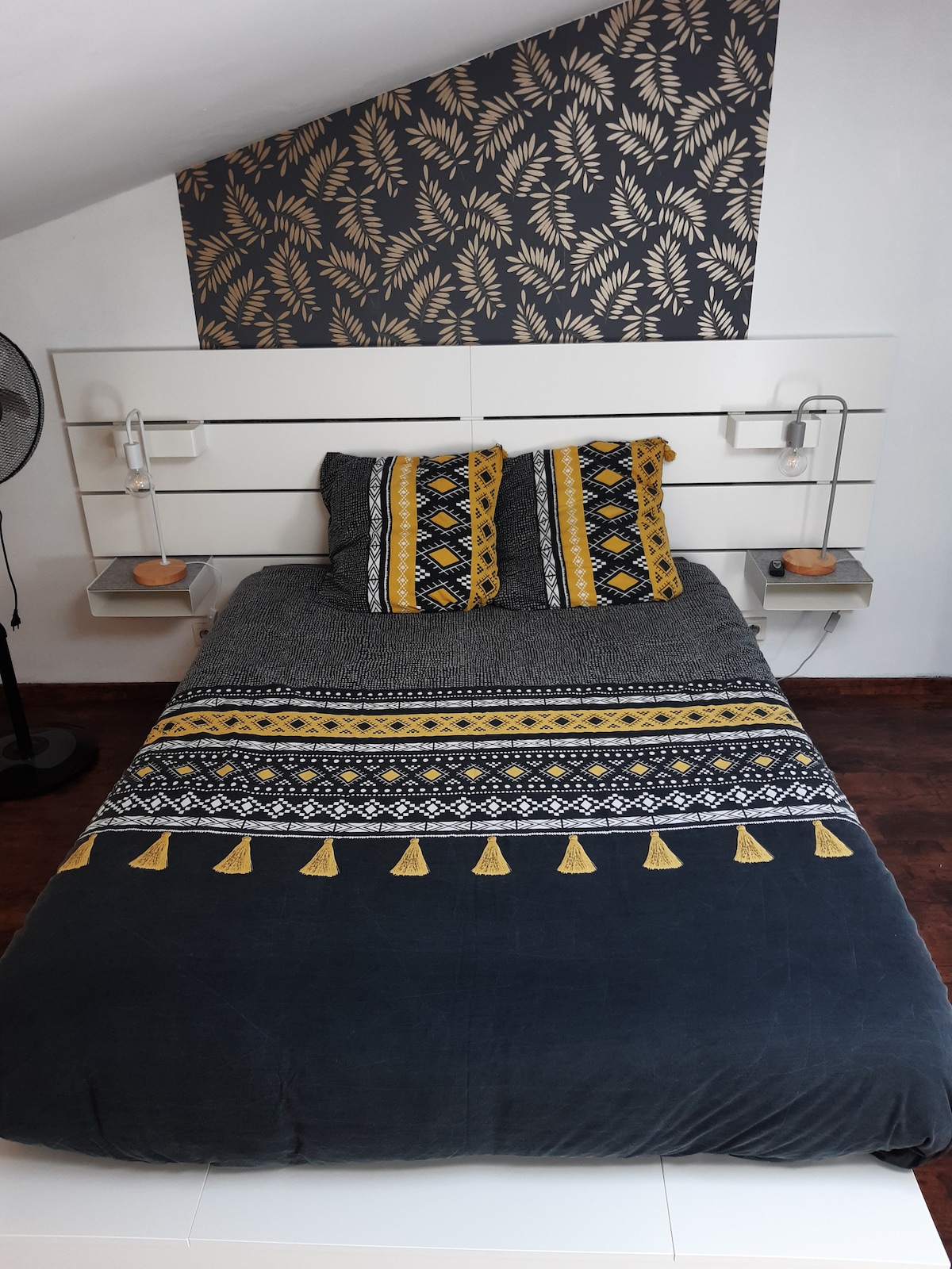 A spacious bed is presented with a decorative duvet featuring intricate patterns in dark tones, complemented by matching pillows. Nightstands are positioned on either side, each holding a small lamp. A stylish leaf-patterned accent wall adds a contemporary touch to the cozy room.
