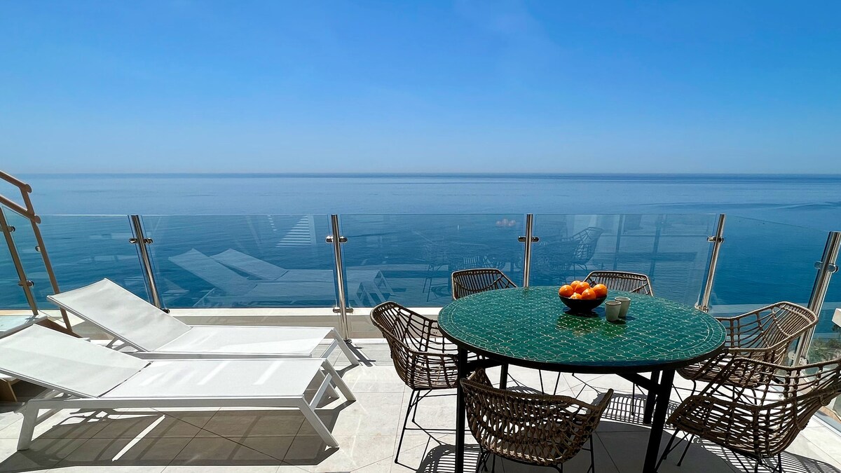 A bright outdoor terrace showcases a circular table surrounded by woven chairs, overlooking a stunning view of the sea. Two sun loungers are positioned nearby, providing a space to relax in the sun, while the clear blue sky reflects on the tranquil water.