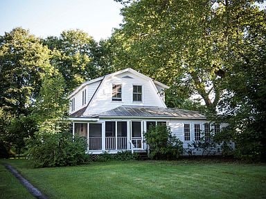 A charming farmhouse is surrounded by lush greenery, featuring a large porch and multiple windows. The exterior is painted white, complemented by a metal roof, and is set on a well-maintained lawn, creating a serene rural setting.