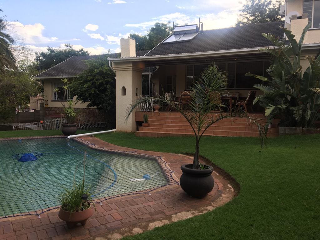 A well-maintained backyard features a sparkling swimming pool surrounded by neatly manicured grass. Potted plants add greenery, while a charming patio area with seating is visible under the rear porch. The scene is set against a backdrop of lush trees and a clear sky.