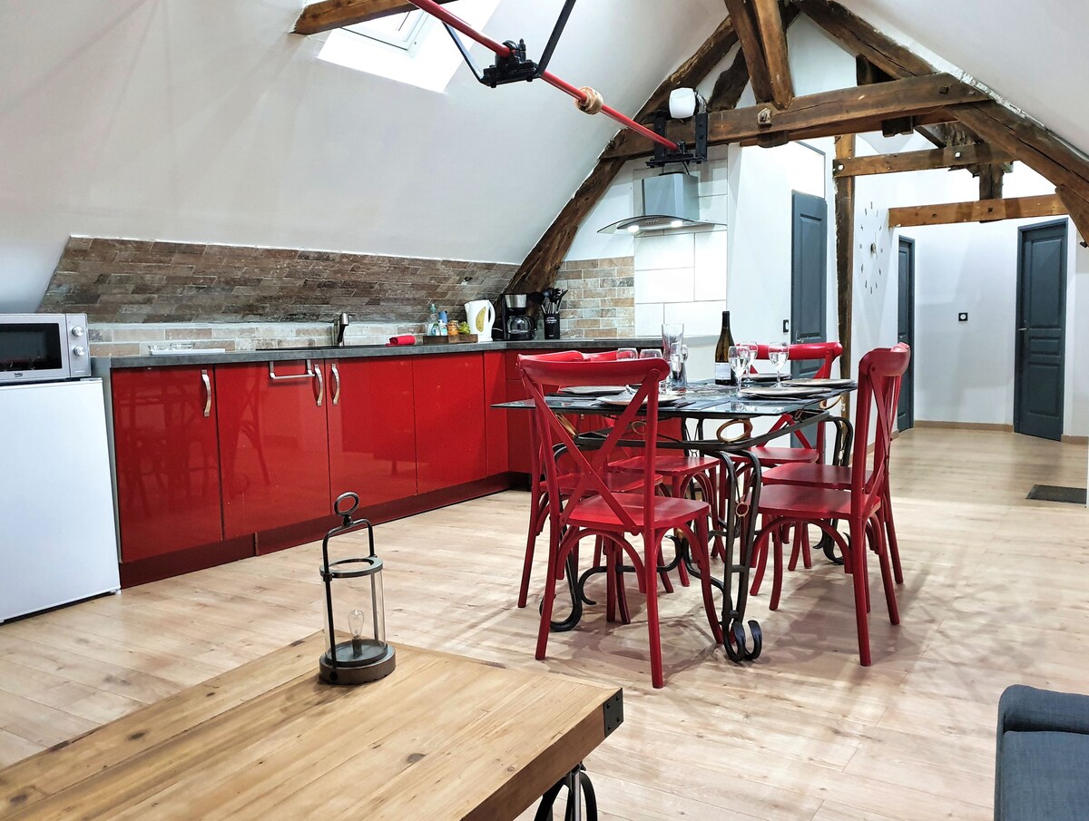 A modern kitchen area showcases bold red cabinetry and sleek appliances. A dining table with red chairs is positioned centrally, complemented by rustic wooden elements. Exposed wooden beams add an architectural detail to the ceiling, while natural light is filtered through a skylight.