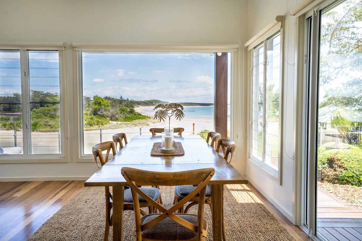 A dining area features a wooden table surrounded by chairs, illuminated by natural light. Large windows provide expansive views of the nearby beach and creek. A neutral rug complements the wooden flooring, creating a relaxed and spacious environment.